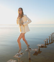 Wearing the Judgement cap toe leather lace-up boots by Bed|Stü, a woman in a light-colored suit stands atop wooden posts in shallow water, with the calm sea and clear sky as her backdrop.