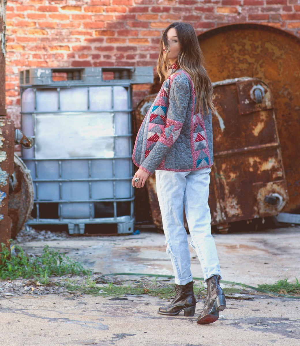 A woman in a patterned jacket, light jeans, and stylish Bed|Stü "Judgement" leather lace-up boots with a block heel stands outdoors on a concrete surface near rusted metal containers and a brick wall.