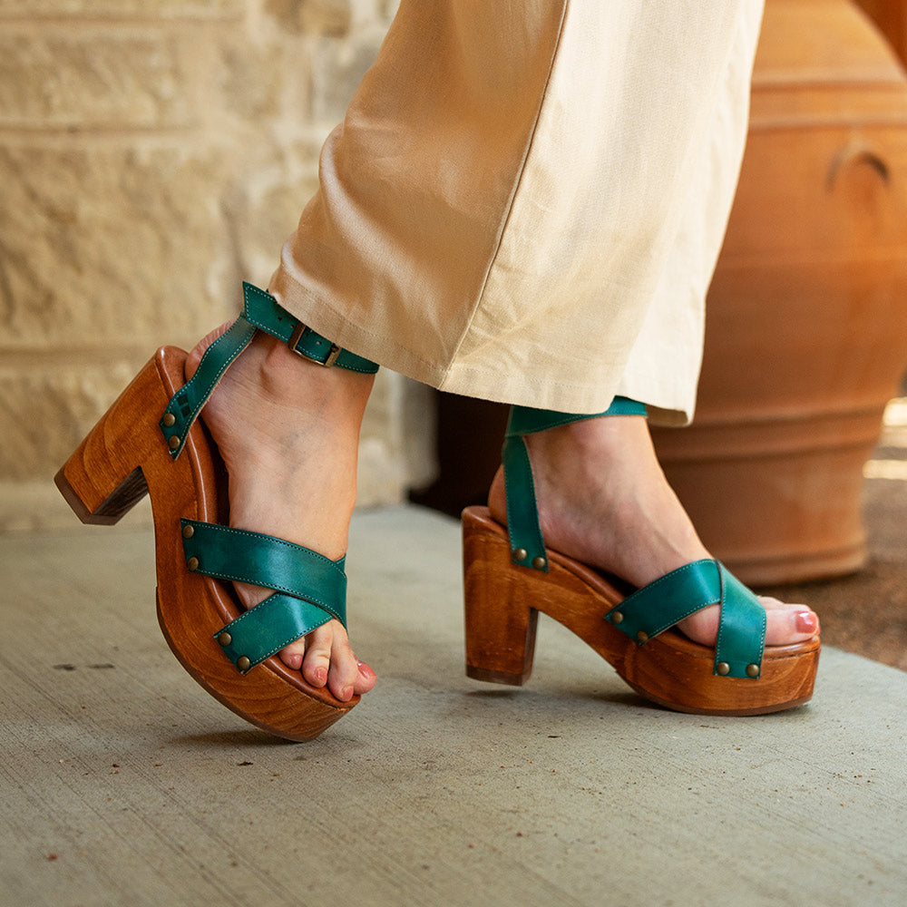 Teal leather platform sandals with crossed straps, ankle buckles, brass-tone studs, and chunky wooden heels, worn with cream trousers.