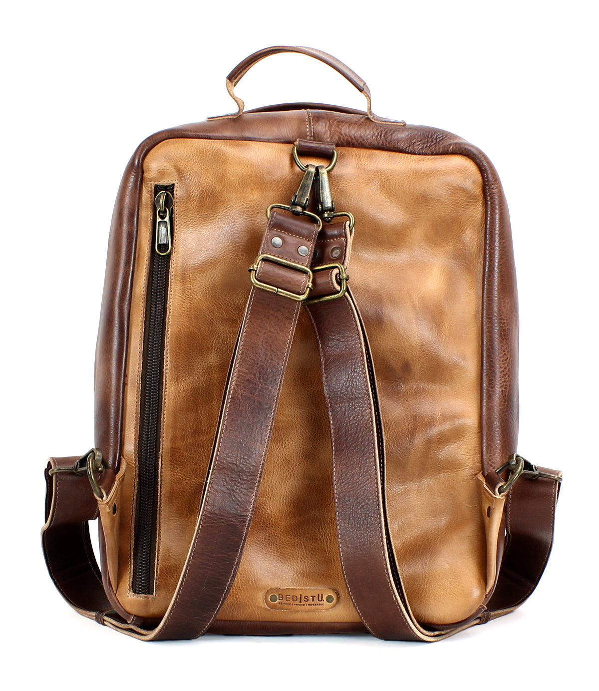 The Bed|Stü Kamet is a brown leather backpack crafted from vegetable-tanned leather, featuring two shoulder straps, a top handle, a side zipper pocket, and brass hardware. This versatile laptop bag is shown against a white background.