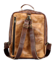 The Bed|Stü Kamet is a brown leather backpack crafted from vegetable-tanned leather, featuring two shoulder straps, a top handle, a side zipper pocket, and brass hardware. This versatile laptop bag is shown against a white background.