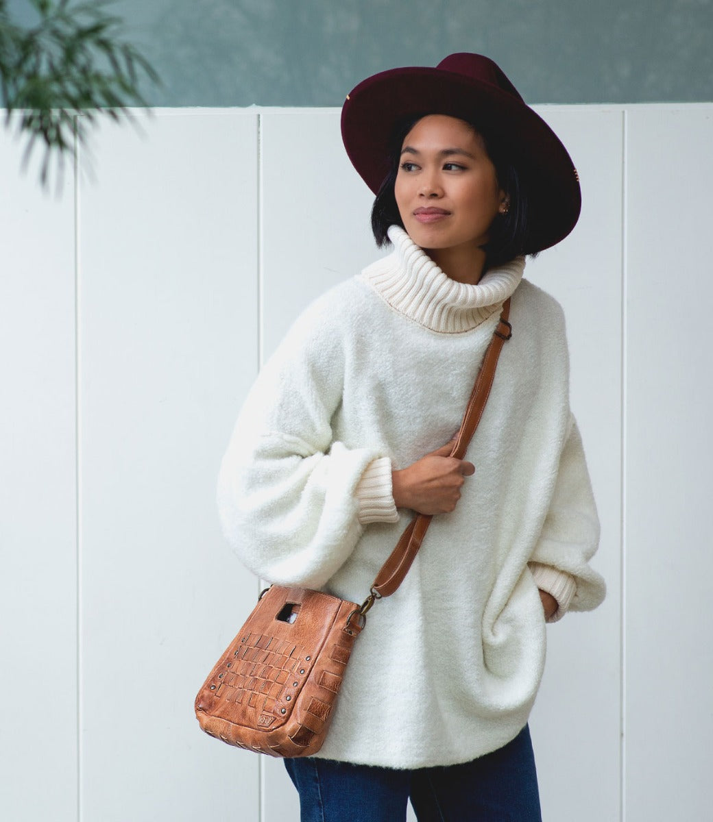 A woman wearing a white turtleneck sweater and maroon hat stands in front of a white fence with her hands in her pockets, carrying the Bed|Stü Keiki checkerboard crossbody bag featuring an adjustable strap.