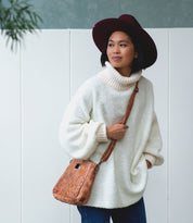 A woman wearing a white turtleneck sweater and maroon hat stands in front of a white fence with her hands in her pockets, carrying the Bed|Stü Keiki checkerboard crossbody bag featuring an adjustable strap.