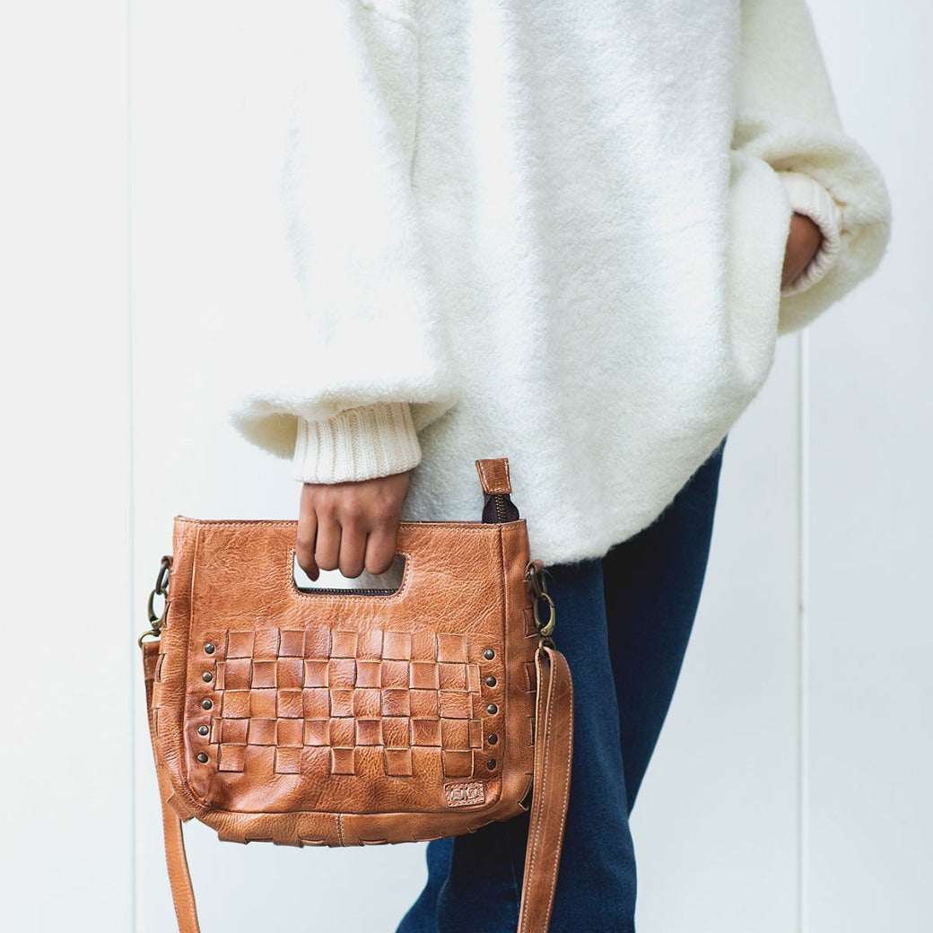 A person wearing a white sweater and blue jeans holds the Bed|Stü Keiki, a tan woven leather handbag featuring metal studs, a zip-top closure, and a shoulder strap.