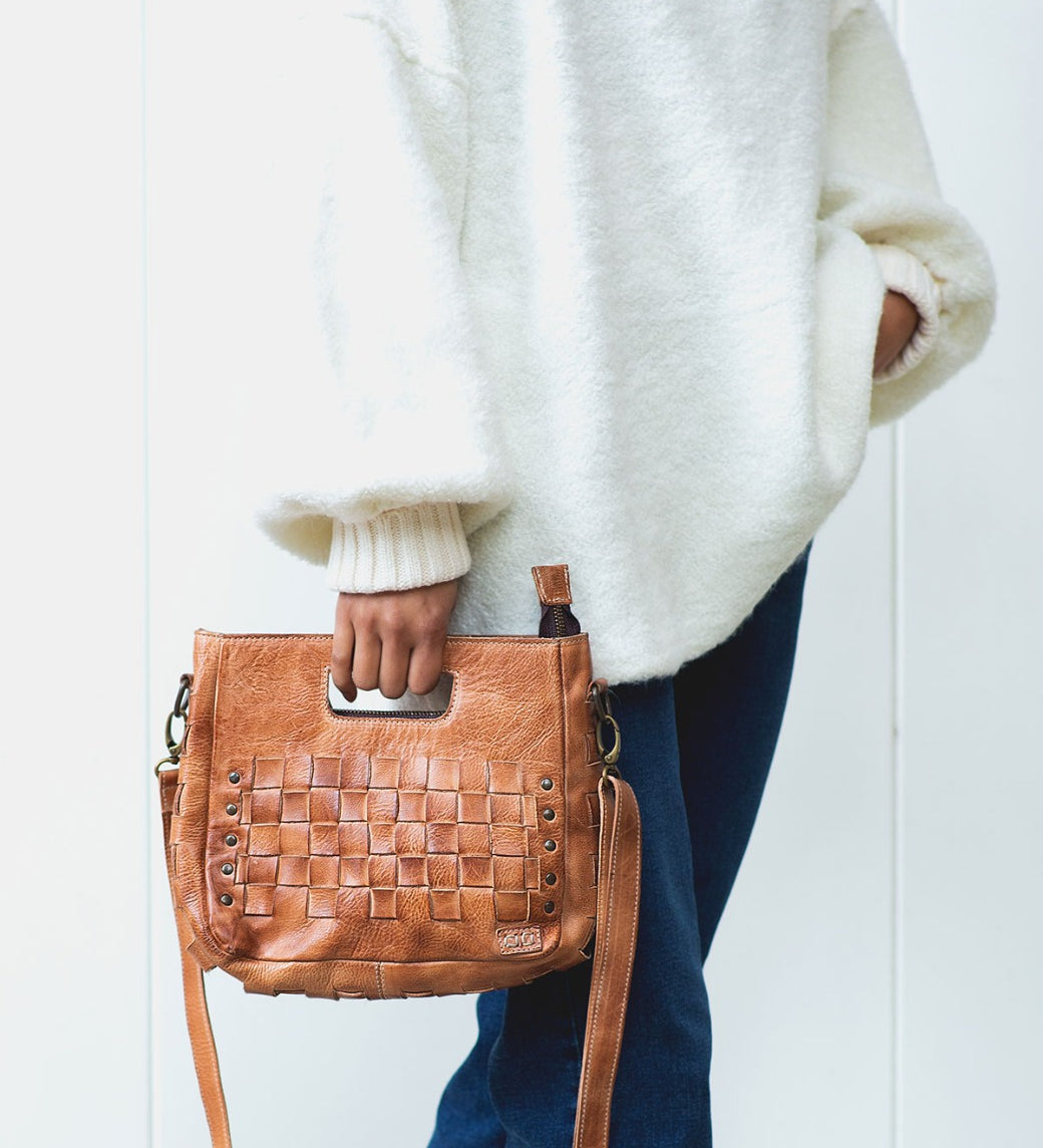 A person wearing a white sweater and blue jeans holds the Bed|Stü Keiki, a tan woven leather handbag featuring metal studs, a zip-top closure, and a shoulder strap.