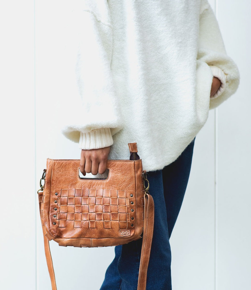 A person wearing a white sweater and blue jeans holds the Bed|Stü Keiki, a tan woven leather handbag featuring metal studs, a zip-top closure, and a shoulder strap.