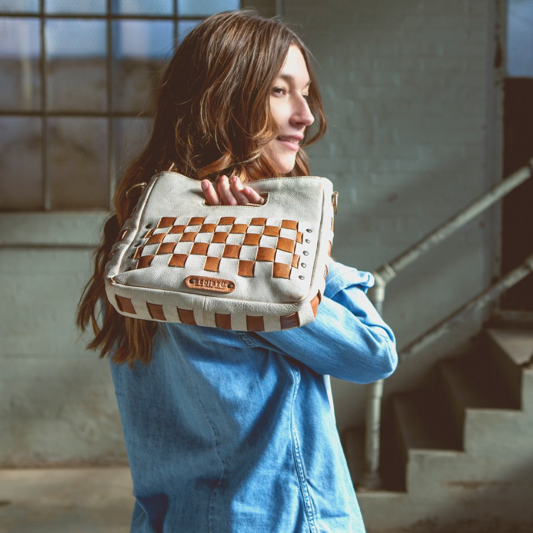 A woman in a denim shirt stands indoors near a window and stairs, carrying the Bed|Stü "Keiki" crossbody bag—a woven beige and brown checkerboard design with an adjustable strap—over her shoulder.
