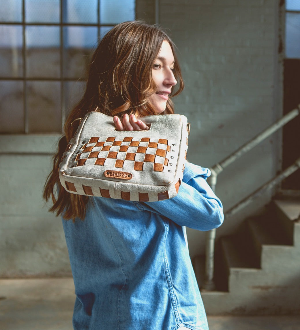 A woman in a denim shirt stands indoors near a window and stairs, carrying the Bed|Stü "Keiki" crossbody bag—a woven beige and brown checkerboard design with an adjustable strap—over her shoulder.