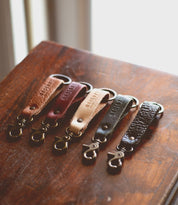 Five Bed|Stü Keygrab full grain leather keychains in assorted brown tones are arranged in a row on a wooden surface, each equipped with a durable swivel clip and metal ring.