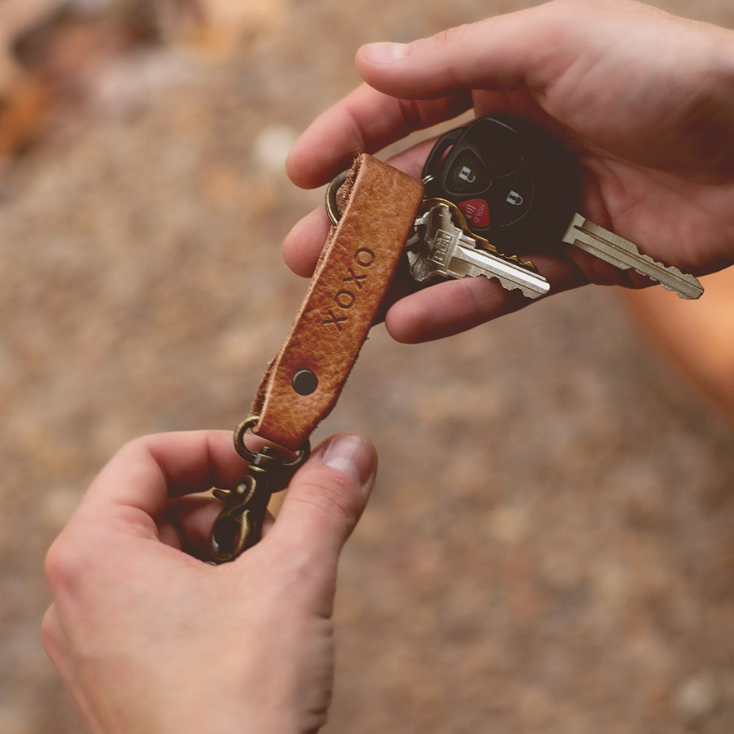 A person holds the Bed|Stü Keygrab—a brown full grain leather strap engraved with "XOXO," featuring a sturdy swivel clip for easy attachment—with two keys and a car remote fob attached.