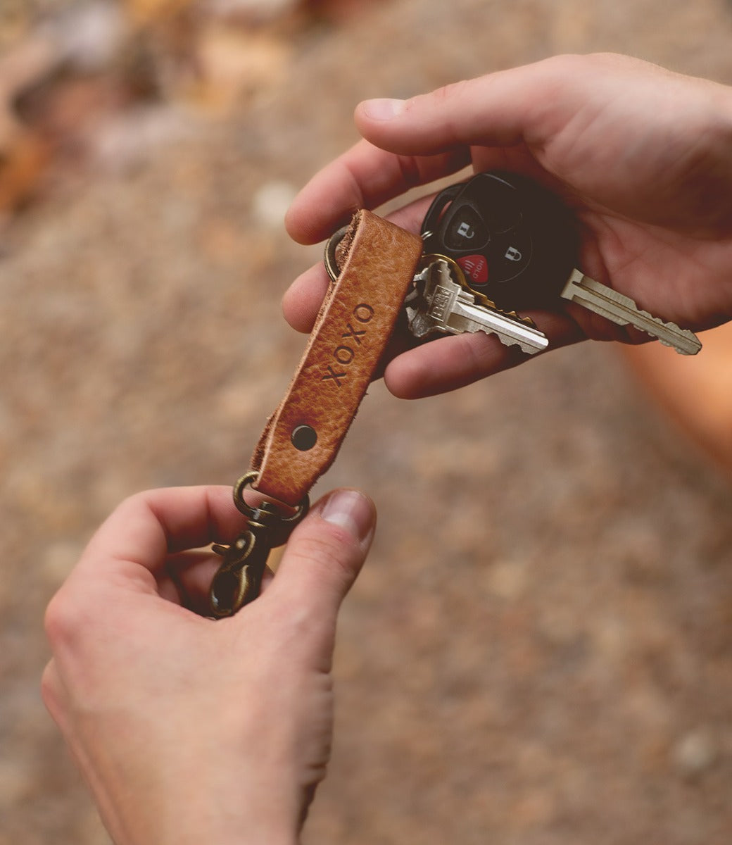 A person holds the Bed|Stü Keygrab—a brown full grain leather strap engraved with "XOXO," featuring a sturdy swivel clip for easy attachment—with two keys and a car remote fob attached.