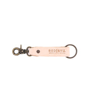 Introducing the Bed|Stü Keygrab: a beige full grain leather keychain with a metal swivel clip and keyring, featuring the Bed|Stü logo and embossed with the words "Genuine | Unique | Imperfect." Perfect as a stylish key organizer.