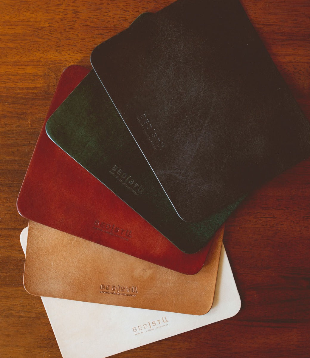 Five rectangular Bed|Stü Launcher Mouse Pads in black, green, brown, tan, and beige are stacked on a wooden surface, each featuring the Bed|Stü logo—making them a stylish addition to any workspace.