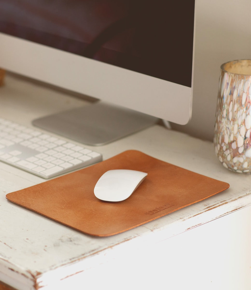 A white computer mouse rests on the Bed|Stü Launcher Mouse Pad, a brown leather workspace accessory, alongside a keyboard, monitor, and speckled cup on a white desk.