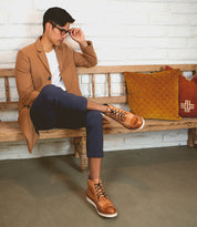 Wearing the Lincoln boots by Bed|Stü, this man in a tan coat, white shirt, and navy pants sits on a wooden bench beside orange and yellow cushions. As he adjusts his glasses, he exemplifies an effortlessly stylish look for everyday wear.