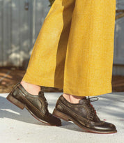 Wearing yellow wide-leg pants and Bed|Stü Lita K II dark brown leather shoes featuring classic derby styling and intricate brogue detailing, the person stands on a light-colored outdoor surface.
