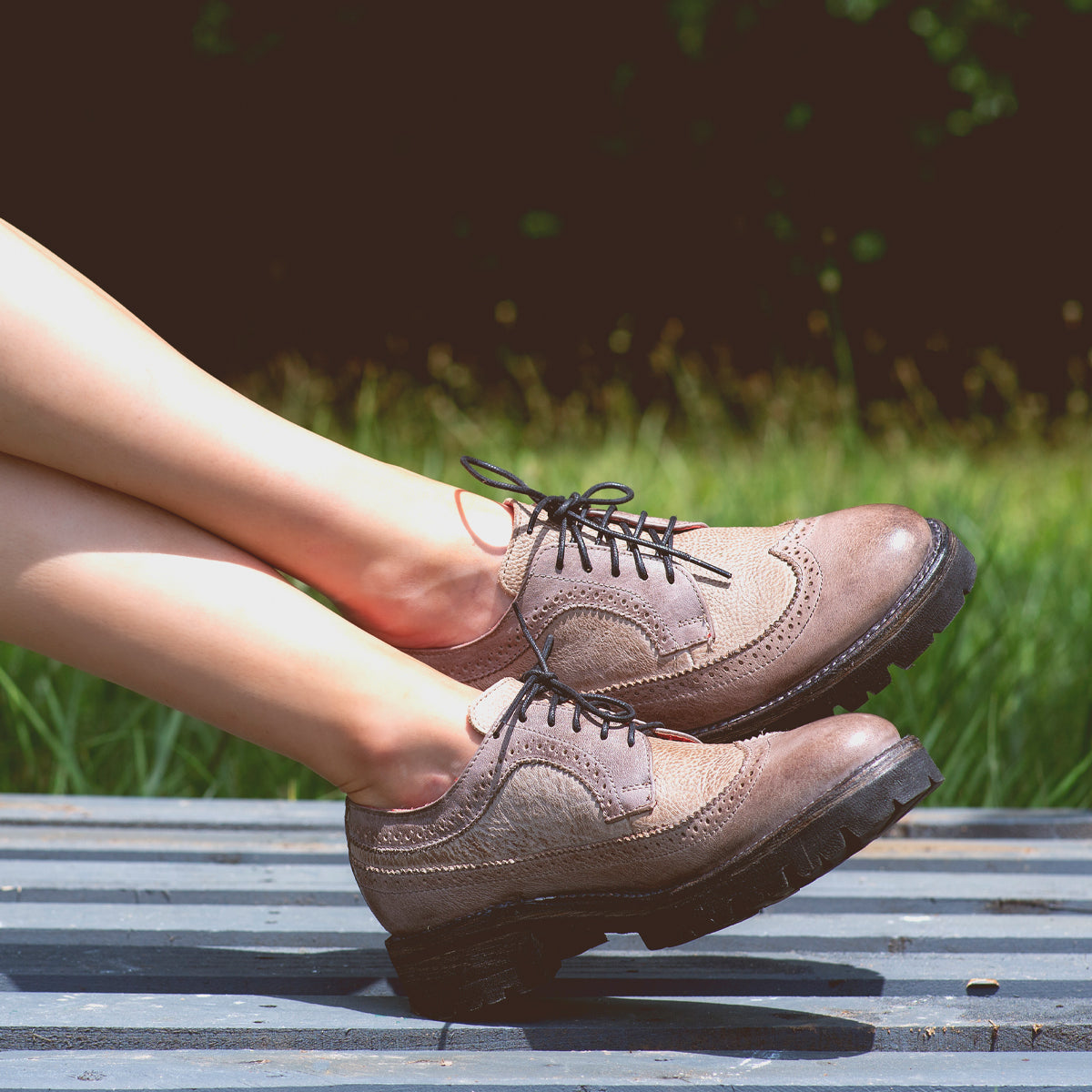 A person with crossed legs wears Bed|Stü Lita K III brown canvas and leather lace-up dress shoes while sitting outdoors on a gray metal surface with grass in the background.