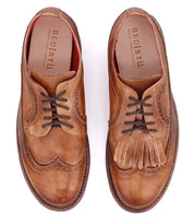 A pair of tan leather dress shoes by Bed|Stü, featuring classic brogue detailing and a wingtip toe; the left shoe has standard laces, while the right showcases Bed|Stü’s Lita K decorative fringe over the laces.