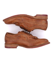 A pair of Bed|Stü Lita K brown leather brogue dress shoes with laces, stacked sole-to-sole, showcasing a classic wingtip toe, intricate brogue detailing, and eye-catching red accents on the soles.