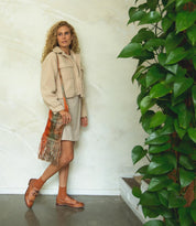 A woman with curly hair in a beige outfit stands indoors beside a green leafy plant, carrying a fringed bag over her shoulder and wearing the Bed|Stü Lita K wingtip toe shoes featuring classic brogue details.
