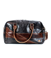 The Luke by Bed|Stü is a black and brown oversized duffle bag crafted from vegetable-tanned leather, featuring dual handles, a shoulder strap, and a small logo patch on the front—perfect as a stylish travel bag.