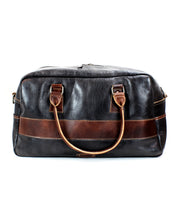 The Bed|Stü Luke, a large black travel bag crafted from vegetable-tanned leather, featuring brown leather handles and a horizontal brown stripe across the front, is displayed against a plain white background.
