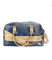 The Luke travel bag by Bed|Stü is an oversized duffle crafted from vegetable tanned leather in blue and tan. Featuring dual handles, a detachable shoulder strap, and a bold diagonal tan stripe, this stylish bag is presented against a white background.