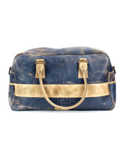 The Luke by Bed|Stü is a blue, oversized duffle bag crafted from vegetable tanned leather, featuring gold-tone handles and a striking gold horizontal stripe, shown against a white background.