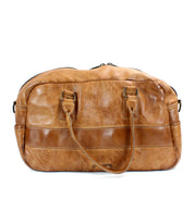 The Luke by Bed|Stü is a medium-sized brown duffel travel bag, expertly crafted from vegetable tanned leather. It features two handles, a front zipper pocket, and side pockets, and is photographed against a white background.