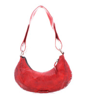 The Bed|Stü Madre is a bold crescent-shaped shoulder bag crafted from red leather, featuring a wide strap adorned with striking metal studs, showcased against a white background for an unmistakable statement look.