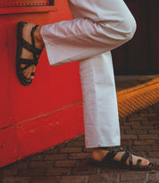 A person wearing white pants and Bed|Stü's handcrafted Manati II leather sandals stands on a brick pavement, one foot resting against a bright red wall.