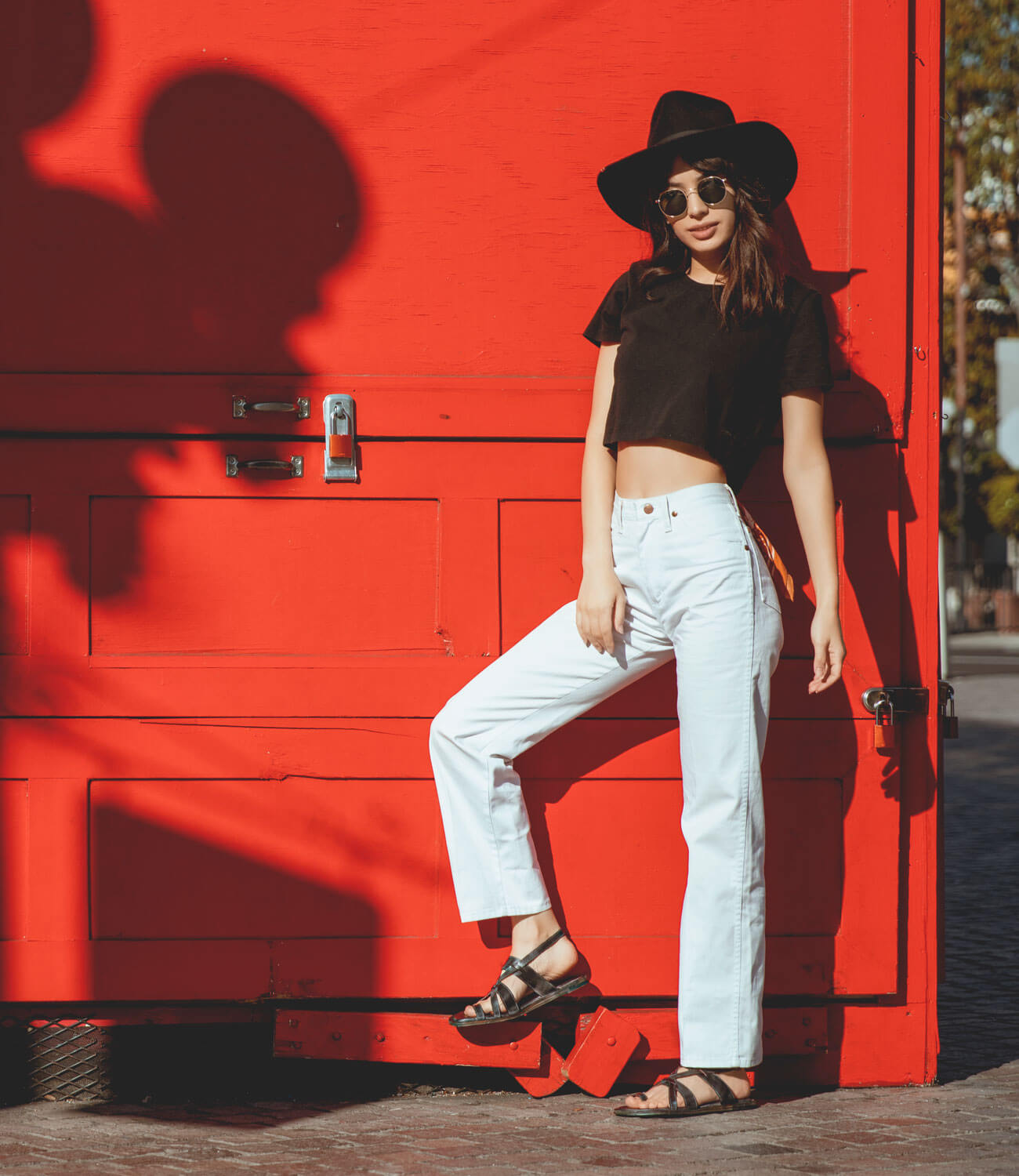 A woman wearing a black hat, sunglasses, black crop top, white pants, and Bed|Stü Manati II handcrafted leather sandals poses against a bright red wall in sunlight.