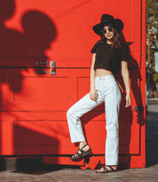 A woman wearing a black hat, sunglasses, black crop top, white pants, and Bed|Stü Manati II handcrafted leather sandals poses against a bright red wall in sunlight.