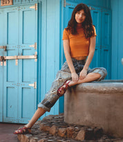 A woman in an orange top and ripped jeans sits on a stone ledge with one leg extended and the other bent, in front of blue doors, highlighting the barefoot comfort of her eco-friendly Bed|Stü Manati II sandals.