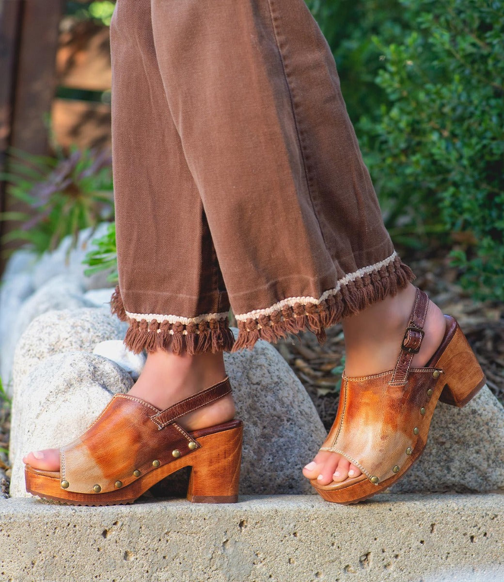 A person wearing brown fringe-hem pants and the Bed|Stü Marie heeled clogs—crafted with vegetable tanned leather uppers and wood platform soles—stands outdoors on a concrete edge with greenery in the background.