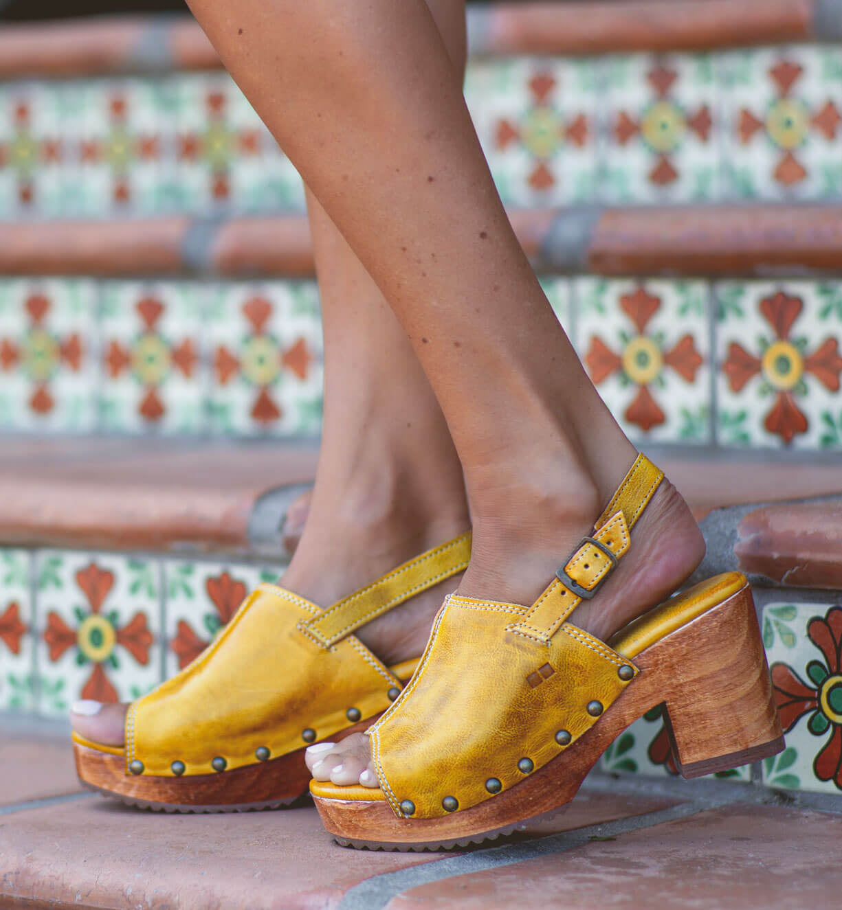 A person wearing the Bed|Stü Marie yellow heeled clogs, crafted from vegetable tanned leather with wooden heels, stands on tiled steps adorned with colorful patterns.