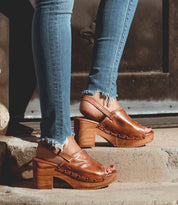 A person wearing frayed hem skinny jeans and the Bed|Stü Marie brown heeled clog sandals, crafted from vegetable tanned leather with wood platform soles and open toes, stands on outdoor steps.
