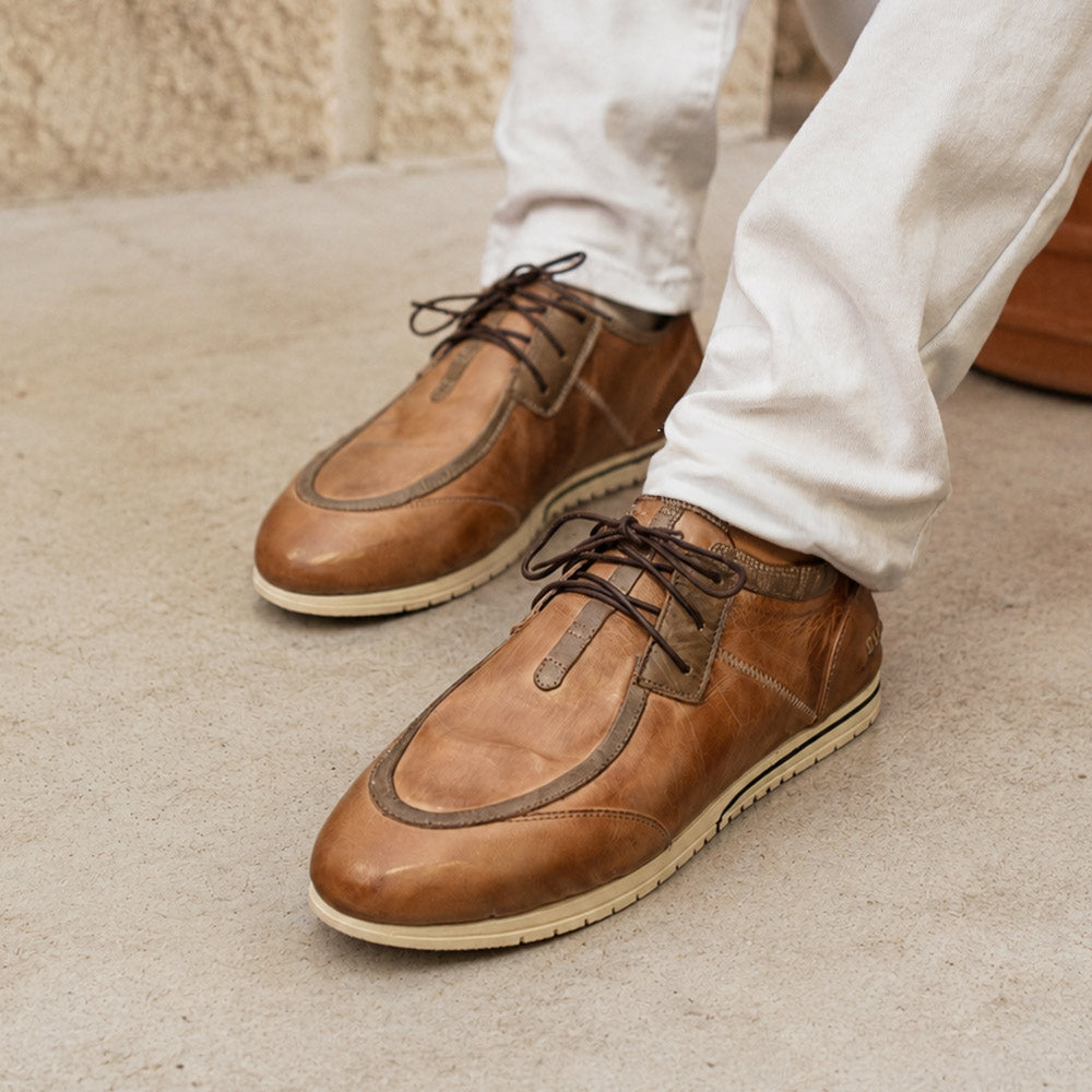 Brown leather lace-up shoes with darker trim and light rubber soles, worn with white trousers on a concrete surface.