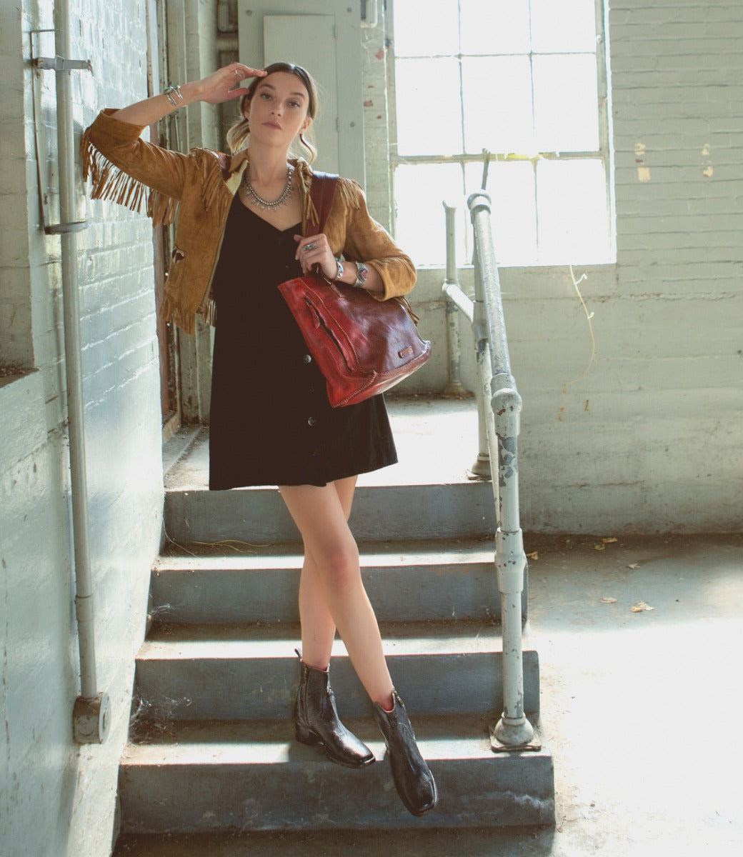 Wearing a fringe jacket, black dress, and hand-stitched leather Bed|Stü boots in the Merryli style, a woman stands on stairs indoors by a window, saluting with her right hand and holding a large brown bag.