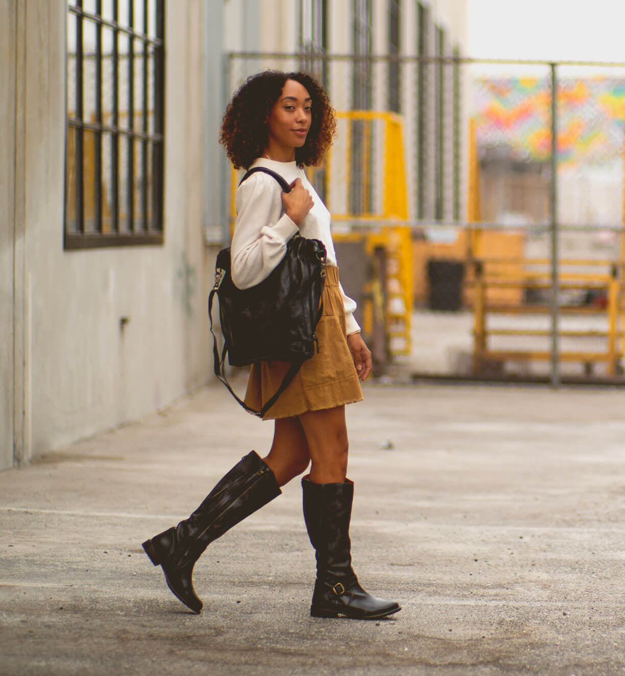 A woman with curly hair walks outside by a fence, wearing a white top, tan skirt, black boots, and carrying the Bed|Stü Mildred—a sleek leather laptop bag with an adjustable strap.