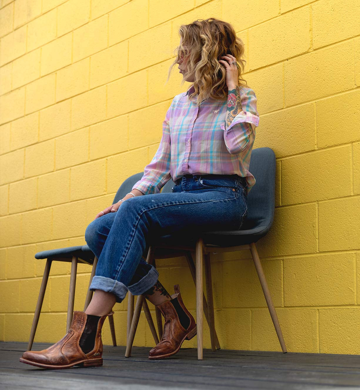 A person sits sideways on a gray chair against a yellow brick wall, wearing a plaid shirt, rolled-up jeans, and the Nandi versatile Chelsea boots by Bed|Stü with a classic slip-on design, with one hand touching their wavy hair.