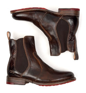 The Nandi boots by Bed|Stü are brown leather Chelsea boots with elastic side panels, pull tabs, and distinctive red rubber soles. They feature a convenient slip-on design, shown with one boot upright and the other positioned on its side.