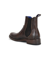 The Bed|Stü Nando is a brown leather Chelsea boot featuring black elastic side panels and a low stacked heel, expertly crafted from hand-worked leather and displayed from a rear side angle on a white background.