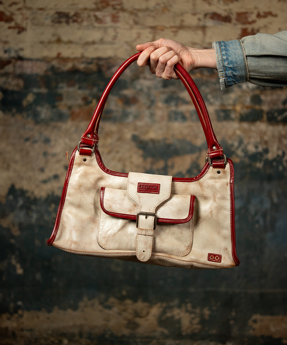 Hand holding a cream leather shoulder bag with red trim, a front buckle pocket, and double handles against a weathered brick wall.