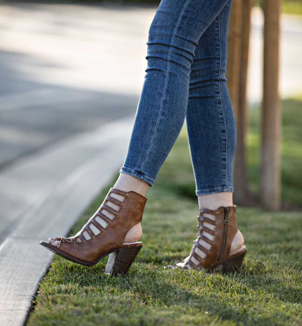 A person wears Bed|Stü's Occam P brown strappy heeled sandals with decorative X stitching, paired with blue skinny jeans, standing with one foot on grass and the other on the curb.