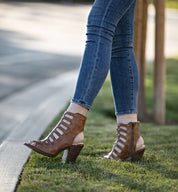 A person wears Bed|Stü's Occam P brown strappy heeled sandals with decorative X stitching, paired with blue skinny jeans, standing with one foot on grass and the other on the curb.