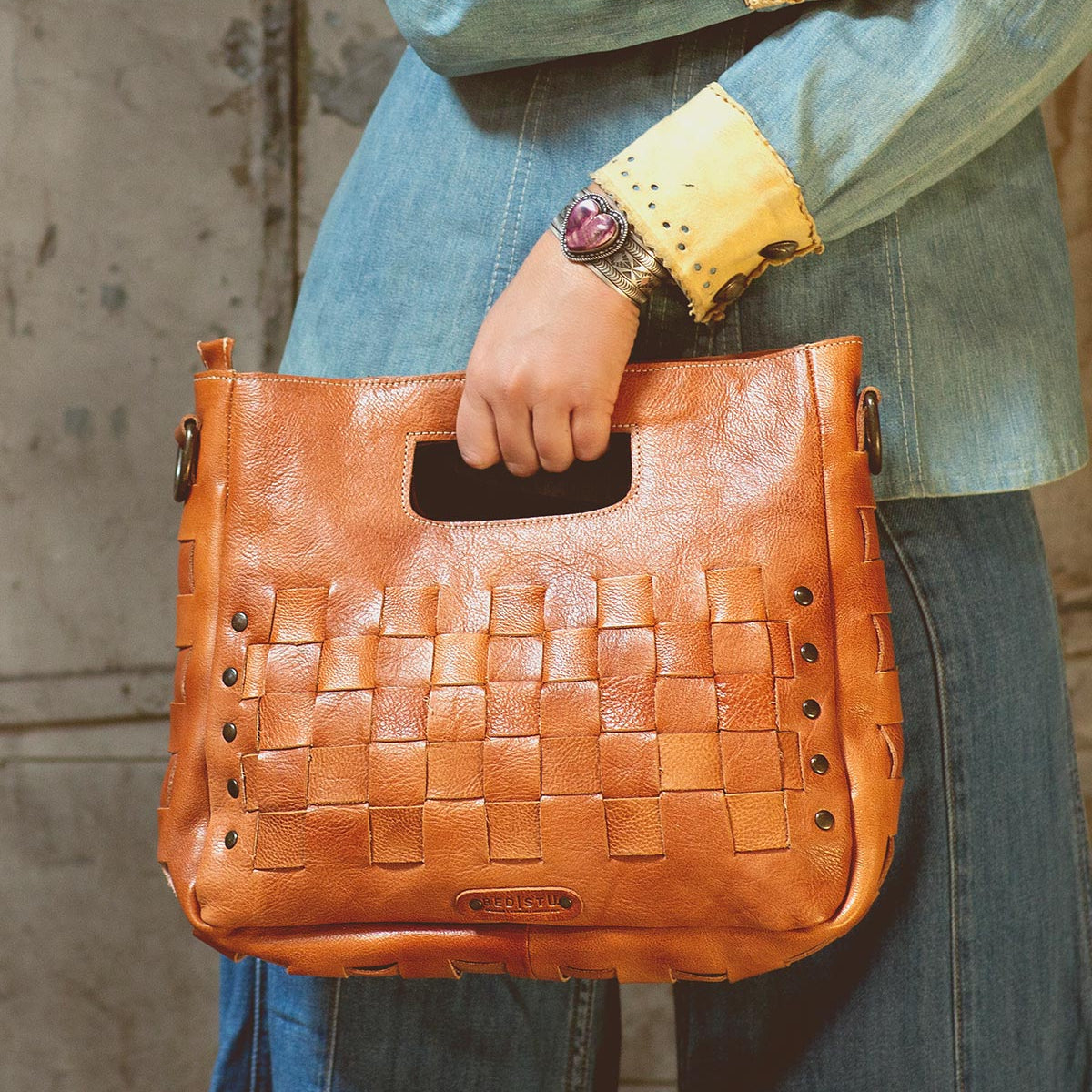 Wearing denim, a person holds the Orchid handwoven leather bag by Bed|Stü, featuring a modern timeless design and a cutout handle, while standing against a textured wall.