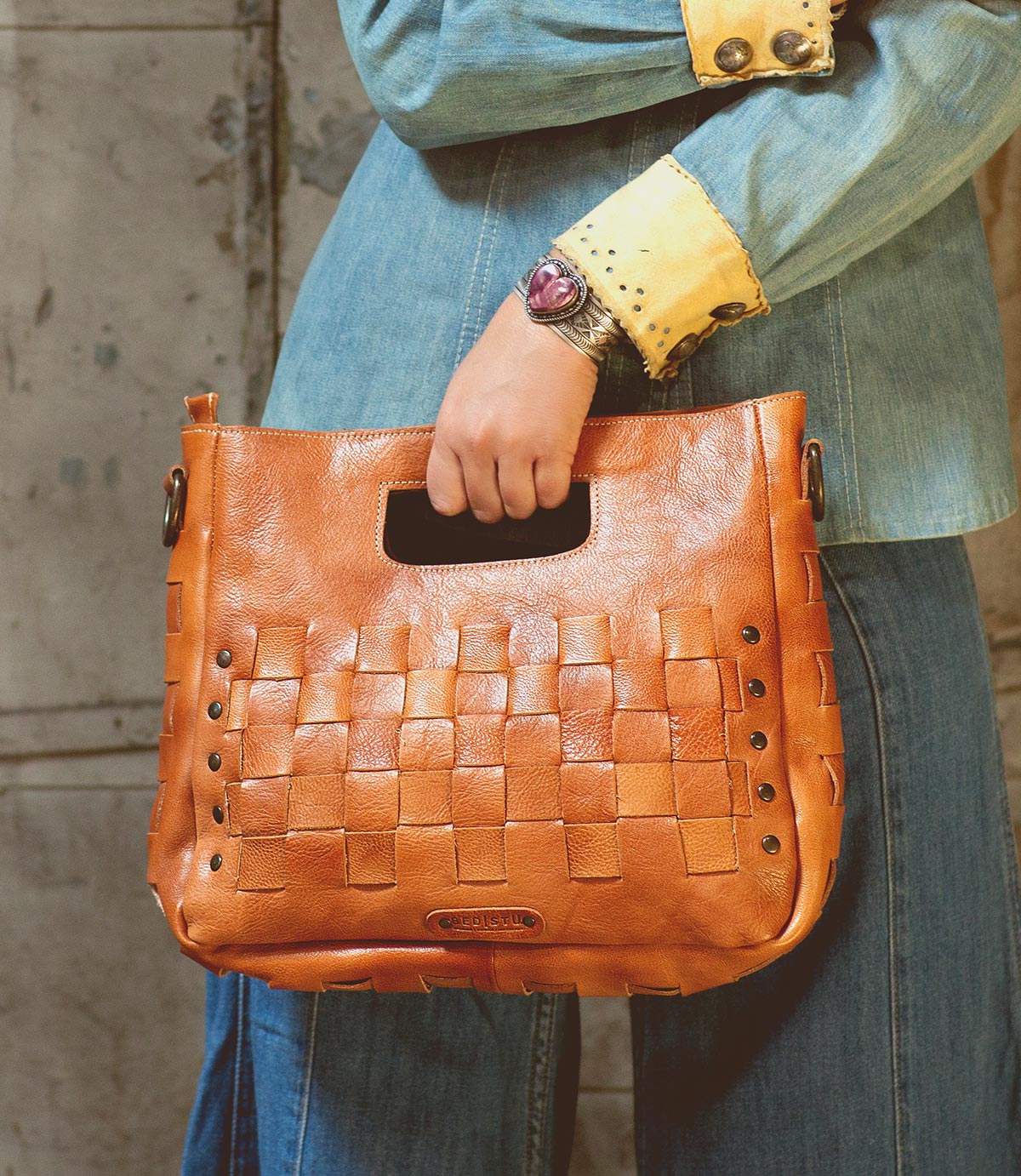 Wearing denim, a person holds the Orchid handwoven leather bag by Bed|Stü, featuring a modern timeless design and a cutout handle, while standing against a textured wall.