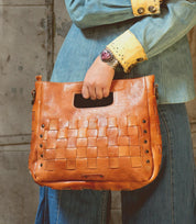Wearing denim, a person holds the Orchid handwoven leather bag by Bed|Stü, featuring a modern timeless design and a cutout handle, while standing against a textured wall.