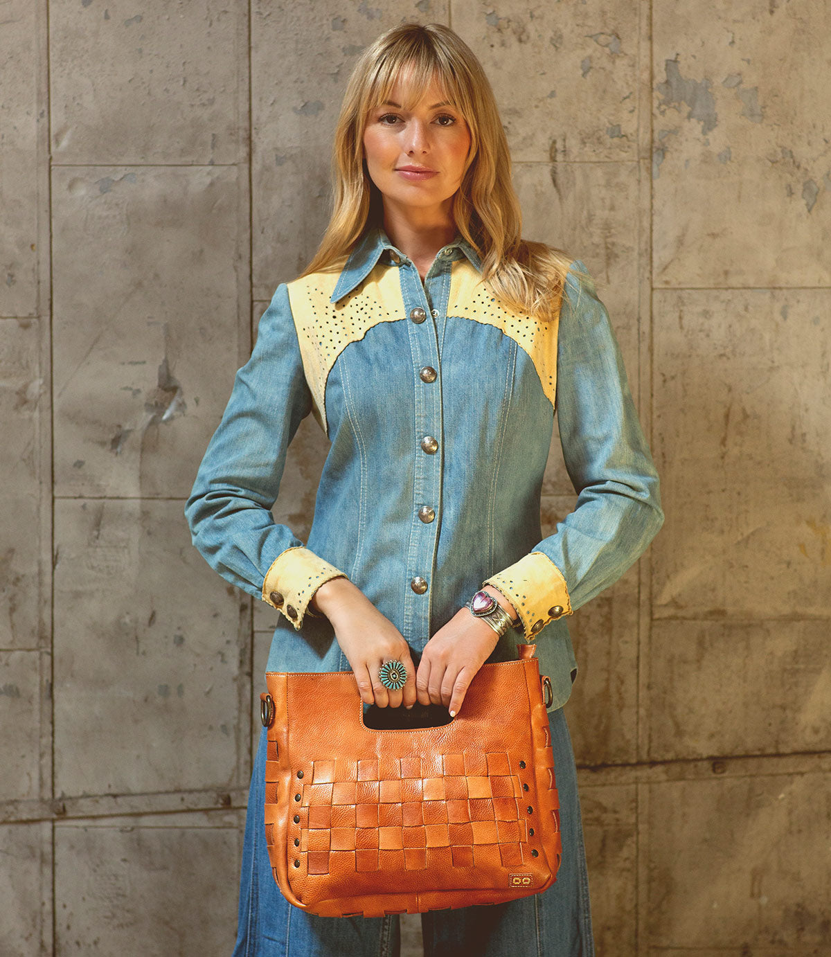 A woman in a denim dress with yellow accents stands against a textured wall, holding the Bed|Stü Orchid handwoven leather bag featuring an antique metal buckle in front of her, effortlessly embodying modern timeless design.